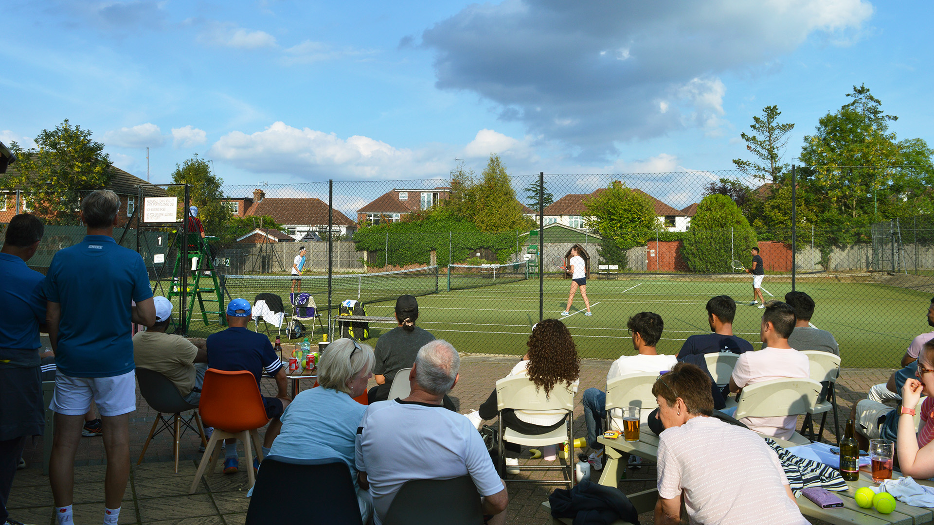 Members and families watching a match from the terrace at Hillfield Close on a summer evening, with players on court